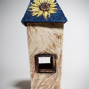 May include: A ceramic house sculpture with a blue roof featuring a yellow sunflower design. The house has a rectangular window and a textured, beige body with brown accents. The artwork is a unique decorative piece.