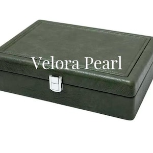 May include: A rectangular dark olive green jewelry box with a textured finish and a silver-toned metal latch closure. The top lid features a stitched border detail, and the white text Velora Pearl is printed across the center of the lid.
