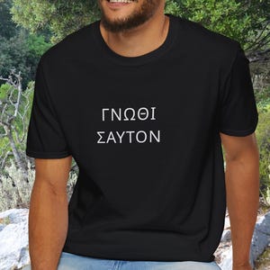 Può includere: T-shirt nero con testo greco bianco. Il testo recita "Conosci te stesso". La maglietta è a girocollo e sembra essere realizzata in un materiale morbido. Lo sfondo è una scena sfocata all'aperto con fogliame verde.