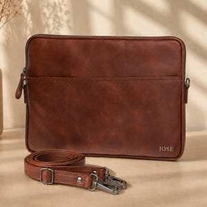May include: A rectangular brown leather laptop sleeve featuring a zippered main compartment, a front slip pocket, and a detachable matching shoulder strap with metal hardware. The name JOSE is embossed in the bottom right corner of the sleeve.