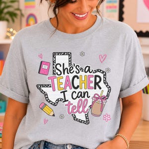May include: A light gray t-shirt featuring a graphic of the state of Texas with a lined paper background. The text reads She's a TEACHER I can tell. The design includes a pink notebook, a yellow pencil, a coffee cup, hearts, and small flowers.