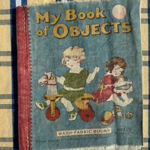 以下が含まれることがあります： 子供と玩具のイラストが描かれた、ヴィンテージの布製本「My Book of Objects」。表紙は色あせた青色で、赤い背表紙があり、「WASH-FABRIC BOOKS」の文字が書かれています。アメリカ合衆国で印刷されました。