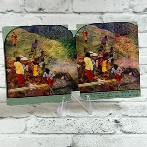 Könnte beinhalten: Eine Vintage-Stereoskopie-Karte mit einer nebeneinander angeordneten, kolorierten Fotografie von Menschen, die in einer Landschaft Wasser schöpfen. Der Text unten rechts lautet: 2. Native Women and Children Fetching Water from a Spring, Malang, Java. Copyrighted, 1903, by T. W. Ingersoll.