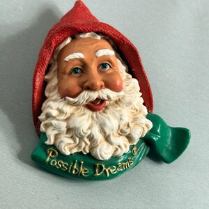 May include: A sculpted Santa Claus face ornament wearing a textured red hood and a green scarf. The figure features a white beard, blue eyes, and a smiling expression. The text Possible Dreams is embossed in gold lettering on the green scarf base.