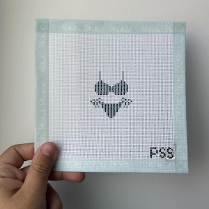 May include: A square needlepoint canvas featuring a centered two-piece bikini design in shades of dark blue and white. The canvas has a light blue fabric border with the text Petite Stitch Studio and the letters PSS stitched in the bottom corner.