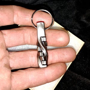 May include: A silver-toned metal keychain featuring a rectangular pendant with a twisted, interwoven design. The pendant is attached to a circular split ring, showcasing a metallic finish with dark, recessed detailing in the grooves.