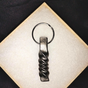 May include: A dark gray metal keychain featuring a vertically oriented, twisted, rectangular bar design. The metal has a textured, forged appearance with a smooth, flat top section where a circular metal split ring is attached for keys.