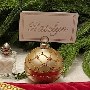 May include: A gold-colored spherical Christmas ornament with a metallic finish, featuring a gold glitter lattice pattern with small red accents. A rectangular place card with the name Katelyn in cursive script is inserted into the top metal cap.