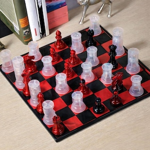 Puede incluir: Un juego de ajedrez con un tablero a cuadros rojo y negro. Las piezas del ajedrez son rojas y negras, con cubiertas de plástico transparente en las piezas blancas. El juego se exhibe sobre una superficie clara.