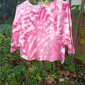 May include: A cropped t-shirt featuring a vibrant pink and white tie-dye spiral pattern. The garment has a crew neckline, short sleeves, and a straight hemline, displayed on a pink plastic hanger against a natural outdoor foliage background.