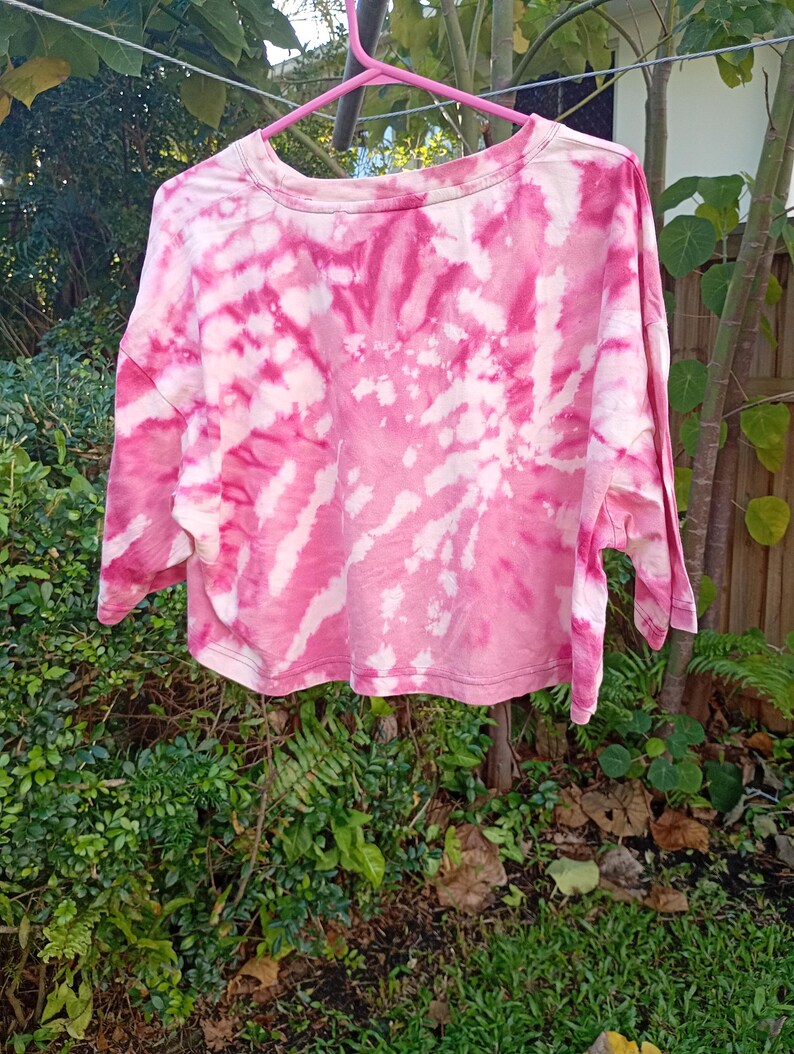 May include: A cropped t-shirt featuring a vibrant pink and white tie-dye spiral pattern. The garment has a crew neckline, short sleeves, and a straight hemline, displayed on a pink plastic hanger against a natural outdoor foliage background.