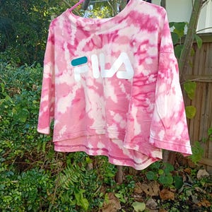 May include: A cropped t-shirt with a pink and white tie-dye pattern featuring the FILA logo printed in white with a teal accent on the letter F. The shirt has a crew neckline, short sleeves, and a high-low hemline with visible stitching.