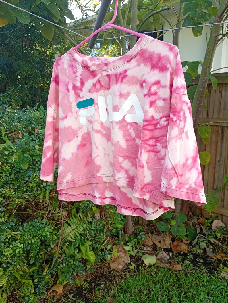 May include: A cropped t-shirt with a pink and white tie-dye pattern featuring the FILA logo printed in white with a teal accent on the letter F. The shirt has a crew neckline, short sleeves, and a high-low hemline with visible stitching.