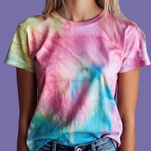 May include: A short-sleeved crew neck t-shirt featuring a tie-dye pattern in shades of pink, blue, and yellow. The fabric displays a swirling, blended colour effect across the front, with a small, subtle embroidered logo detail on the right sleeve.