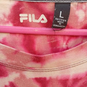 May include: A Fila brand t-shirt with a pink and white tie-dye pattern. The interior neck label displays the Fila logo, size L, and manufacturing details including RN129164 and Made in Bangladesh. The shirt is draped over a pink hanger.