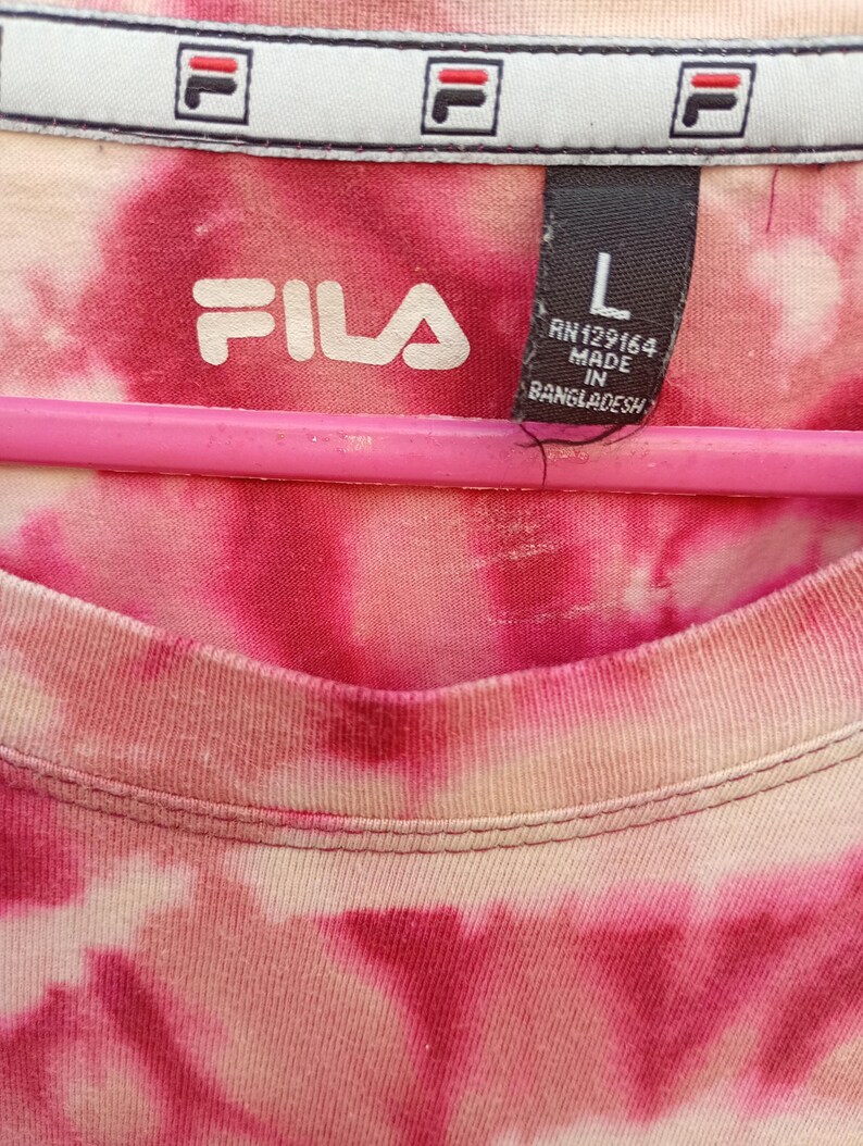 May include: A Fila brand t-shirt with a pink and white tie-dye pattern. The interior neck label displays the Fila logo, size L, and manufacturing details including RN129164 and Made in Bangladesh. The shirt is draped over a pink hanger.