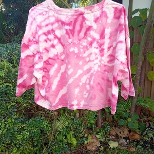 May include: A cropped t-shirt featuring a pink and white tie-dye spiral pattern. The garment has a crew neckline, short sleeves, and a straight hemline. The fabric displays a swirling design of vibrant pink hues contrasted with white areas.