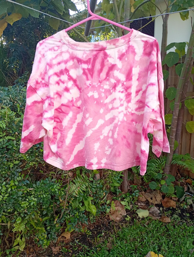 May include: A cropped t-shirt featuring a pink and white tie-dye spiral pattern. The garment has a crew neckline, short sleeves, and a straight hemline. The fabric displays a swirling design of vibrant pink hues contrasted with white areas.