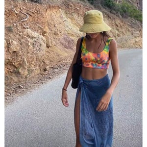 Puede incluir: Top corto con estampado tie-dye en tonos naranja, amarillo y morado, combinado con un pareo azul y un sombrero de pescador amarillo. El conjunto se completa con un bolso tote negro y una pulsera negra. La persona camina por una carretera asfaltada.