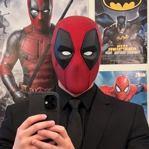 Może przedstawiać: Czerwono-czarna maska Deadpoola jest noszona z czarnym garniturem i krawatem. Osoba trzyma czarny telefon, robiąc sobie selfie w lustrze. Plakaty z superbohaterami są widoczne w tle.