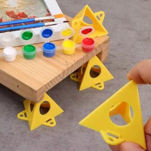 May include: A wooden artist's palette with paint pots in various colors, including white, green, blue, yellow, and red. Paintbrushes and yellow triangular palette supports are also visible, suggesting a painting kit.