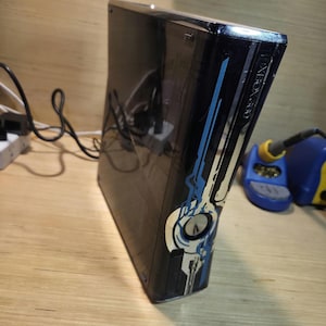 May include: Black Xbox 360 console with blue and white decorative accents. The console is on a wooden surface. The text "XBOX 360" is visible on the side of the console.