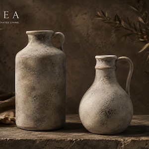 Puede incluir: Dos jarras de cerámica con un acabado texturizado y desgastado en tonos beige y gris. La jarra más alta es cilíndrica, mientras que la más corta tiene un cuerpo redondeado. Ambas tienen un asa lateral y el texto BOVEA TIMELESS DECOR, ELEVATED LIVING.