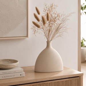 Può includere: Un vaso in ceramica beige con base ampia e collo stretto contiene erbe ornamentali essiccate. Il vaso è posto su un mobile in legno chiaro, con una ciotola e dei libri. L'estetica generale è minimalista e neutra.