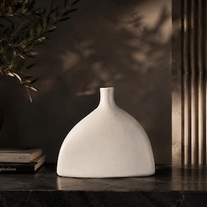 Peut inclure: Un vase en céramique blanc mat avec une base large et arrondie et un col étroit centré. Le vase présente une silhouette lisse et incurvée qui s'effile vers le haut jusqu'à une petite ouverture circulaire, offrant une surface unie.