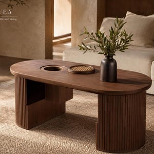 Peut inclure: Une table basse en bois foncé avec un plateau ovale lisse, dotée d'une découpe circulaire et d'un petit plateau rond tressé. La table possède deux supports incurvés à lattes verticales. Un vase noir mat avec du feuillage vert est posé dessus. Texte : BOVEA.