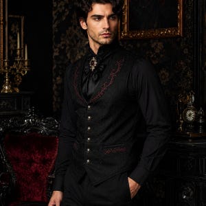 Könnte beinhalten: Eine schwarze Weste mit dezentem Strukturmuster, einer Reihe silberfarbener Knöpfe und dunkelroter Stickerei an den Revers und Taschen. Die Weste wird über einem schwarzen Langarmhemd mit einem schwarzen Plastron und einer dekorativen Brosche getragen.
