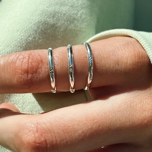 May include: Three silver rings with a simple geometric design are worn on a finger. The rings are thin bands with a slightly curved shape. The rings are made of a shiny metal, reflecting the light. The rings are a minimalist jewelry item.