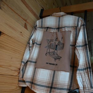May include: A long-sleeve button-down shirt in a beige, white, and dark brown plaid pattern. A large, square brown fabric patch is sewn onto the back, featuring a black line-art graphic of a skeleton in a shopping cart with the text LIVE YOUR BEST LIFE.