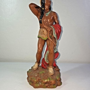 Puede incluir: Figura de nativo americano de piel morena, de pie sobre una base marrón y verde. La figura lleva un taparrabos beige, mocasines y un chal rojo y verde. Una pluma adorna la cabeza de la figura. Un artículo decorativo.