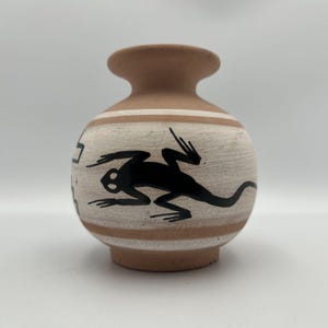 May include: A small terracotta vase with a rounded body and a narrow neck. The surface features horizontal bands in white and natural clay tones, decorated with a black silhouette of a lizard and geometric patterns painted around the center.