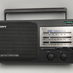 May include: A black Sony portable radio with a top handle, featuring a horizontal speaker grille on the left and a silver tuning dial panel on the right. The display shows AM, FM, TV, and weather bands with printed text including SONY and ICP-54.