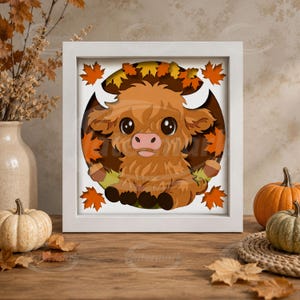 May include: A square white shadow box frame containing a layered paper art cutout of a fluffy brown Highland cow. The cow is centered within a circular dark brown background, surrounded by orange and yellow autumn maple leaves and small brown acorns.