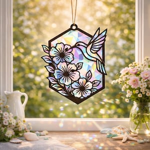 May include: A decorative hummingbird and floral suncatcher. The suncatcher is a dark brown hexagon shape with a hummingbird and flowers cut out. The center is filled with iridescent glass. The suncatcher hangs from a gold chain.