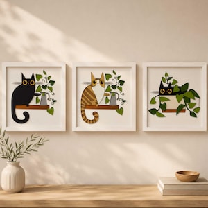 Könnte beinhalten: Drei gerahmte Kunstdrucke mit Katzenmotiven. Jeder Druck zeigt ein anderes Katzendesign, darunter eine schwarze Katze, eine getigerte Katze und eine schwarze Katze zwischen Blättern. Die Katzen sitzen auf einem Holzregal mit grünen Blättern und einer kleinen Gießkanne.