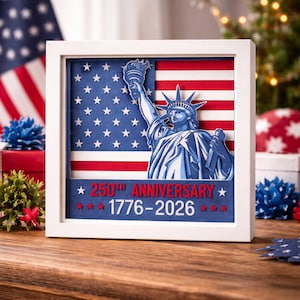 Könnte beinhalten: Ein gerahmtes Papierkunstwerk mit der Freiheitsstatue vor dem Hintergrund der amerikanischen Flagge. Das Kunstwerk ist weiß gerahmt und enthält den Text "250th Anniversary 1776-2026" in Rot und Weiß.