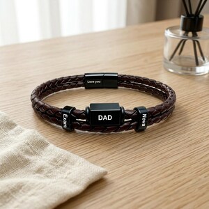 Puede incluir: Una pulsera de cuero trenzado marrón oscuro con un cierre de metal negro grabado con el texto Love you. La pulsera incluye tres cuentas de metal negro, con la cuenta central mostrando la palabra DAD y las dos laterales mostrando Exam y Nova.