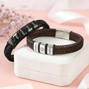 Op de afbeelding: Twee gepersonaliseerde leren armbanden. Eén is zwart met zilveren naamkralen, de andere is bruin met zilveren kralen en een smiley-kralen. Beide armbanden hebben een gevlochten ontwerp en een metalen sluiting.