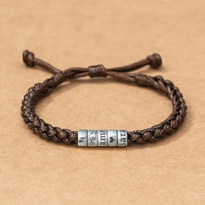Puede incluir: Una pulsera de cordón trenzado marrón con cinco cuentas cilíndricas de tono plateado. Las cuentas están grabadas con una figura de palo, un gesto de promesa, el texto "ame", un símbolo de corazón y el texto "Sara" en fuente oscura.