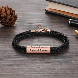 May include: Black braided bracelet with rose gold accents and clasp. The bracelet features two rose gold bars engraved with the words "Forever & Always" and the names "Fallon & Raven".