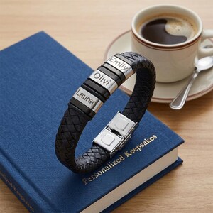 May include: A black braided leather bracelet featuring three rectangular silver-toned metal beads. Each bead is engraved with a name: Emily, Olivi, and Lauren. The bracelet includes a polished silver-toned metal clasp mechanism for secure closure.