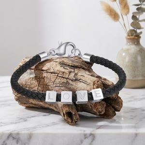 May include: A black braided leather bracelet featuring four silver-toned square beads, each engraved with a letter to spell the word LOVE. The bracelet is secured with a silver-toned lobster claw clasp and metal end caps on both sides of the cord.
