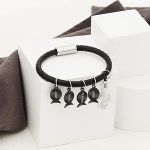 May include: Black braided leather bracelet with a silver clasp. The bracelet features four black charms shaped like fish, each with a name engraved, and a silver fish hook charm. The names are Jonah, Simon, Solomon, and Josiah.