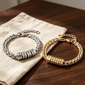 May include: Two wheat chain bracelets, one silver-toned and one gold-toned, each featuring four cylindrical beads engraved with names including Pippa, Charlie, and Halle. Both bracelets have a lobster claw clasp and an adjustable extender chain.