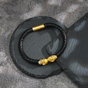 May include: Black braided leather bracelet with gold-colored skull accents. The bracelet features a gold-colored clasp. The skulls are detailed with intricate carvings. The bracelet is displayed on a dark gray, textured surface.