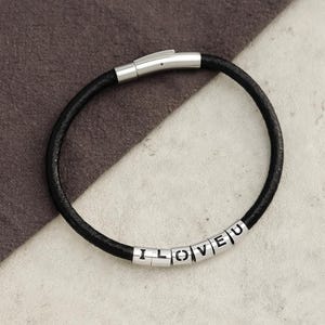 May include: Black leather bracelet with silver-tone clasp and beads spelling "I LOVE U". The bracelet is circular and appears to be made of braided leather. The beads are small and rectangular.
